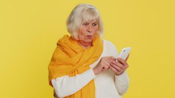 Upset sad senior woman use smartphone typing browsing, play game loses surprised by sudden lottery results bad fortune loss fail unlucky news. Elderly mature pensioner grandmother on yellow background video