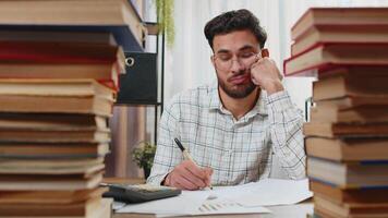 Exhausted Indian businessman reviewing documents reports while using calculator working hard in home office. Stressed alone freelancer touching head in despair surrounded by stacks of books on table. video