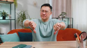 Dislike. Upset unhappy middle-aged Chinese man showing thumbs down sign gesture, expressing discontent disapproval dissatisfied bad work at home apartment indoors. Displeased Asian guy in room on sofa video