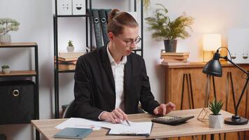 Focused young office worker working with schedules and financial graphics while using calculator sitting at home office desk. Happy stylish businessman freelancer compare and analyze graphs charts. video