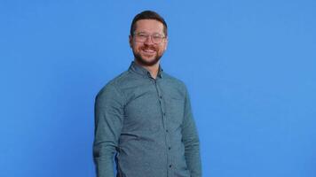 Hey you. Happy Caucasian young man in glasses smiling excitedly and pointing forward to camera, choosing lucky game winner, indicating to awesome you, promotion. Guy isolated on blue background video