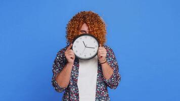 Serious strict man with curly hairstyle wig holding office clock hiding behind, checking time on watch, running late to work, being in delay, deadline, obscuring face. Guy isolated on blue background video
