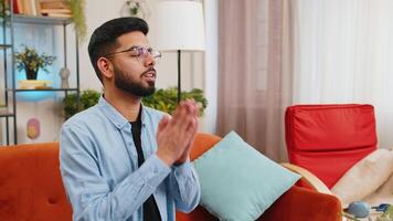 Tensed Hispanic man sits on sofa at home folding palms together pray ask for help thinks feel stressed looks desperate by life problems. Worried upward Indian guy praying making wish begging apology video