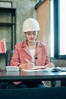 Female engineer wearing a hard hat, working diligently at a desk in a modern office setting. photo