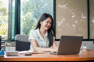 Smiling young woman using a laptop at a desk in a bright, modern office with large windows and greenery outside. photo