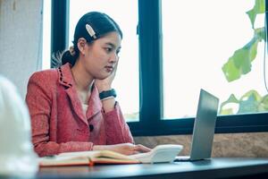 Young woman in a red blazer working on a laptop at a desk, with natural light streaming through a window in a modern office. photo