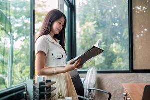 A young woman stands in a modern office, reviewing documents. Large windows provide ample natural light and a view of greenery. photo