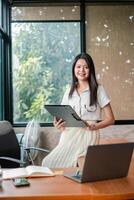 Confident young woman in a modern office setting, holding a tablet and smiling, with a laptop and large windows in the background. photo