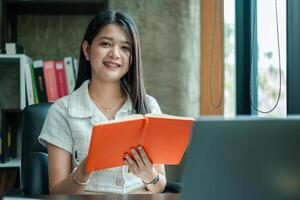 A young woman sits at a desk in a modern office, reading a book. Natural light streams through a window, creating a serene atmosphere. photo
