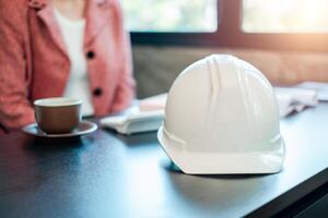 White hard hat on a desk with a businesswoman in the background, representing construction and engineering concepts. photo