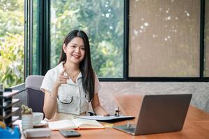 Smiling woman in a modern office setting, working on a laptop with documents and a calculator on the desk. photo