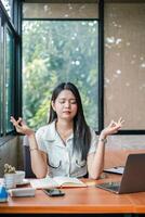 A young woman practices meditation at her office desk, surrounded by a laptop and notebook, in a bright and modern workspace. photo