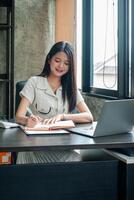 A young woman sits at a desk, writing in a notebook with a laptop open, in a bright and modern home office setting. photo