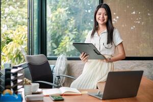 Smiling young woman in a modern office holding a tablet, with a laptop and documents on the desk, surrounded by greenery. photo