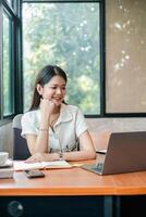 Smiling young woman using a laptop at a desk in a bright, modern home office setting with natural light. photo