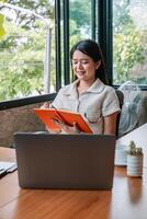 A young woman working on a laptop and taking notes in a bright, modern office with large windows and greenery outside. photo