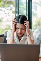Woman feeling stressed while working on a laptop in a home office setting with natural light and greenery visible through the window. photo