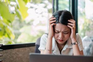 A woman appears stressed while working on a laptop in a modern office setting with natural light and greenery outside. photo