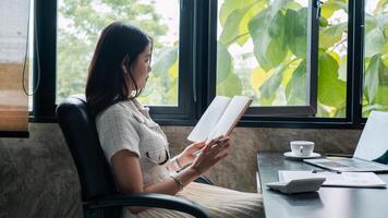 A woman sits by a window reading a book in a modern office, surrounded by greenery and natural light. photo