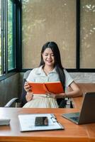 A young woman sits at a desk in a modern office, smiling while reading a tablet. Natural light filters through large windows. photo