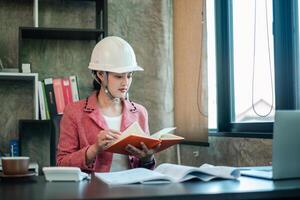 Female architect wearing a hard hat, reading plans at a modern office desk with natural light streaming through the window. photo