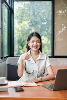 Confident young woman in office, smiling and giving thumbs up, with laptop and notebook on desk. photo