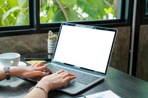 Hands typing on a laptop with a blank screen in a cozy cafe, surrounded by plants and natural light. photo