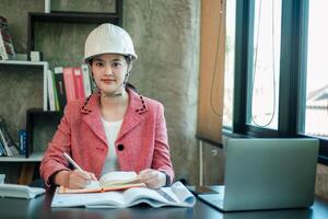 Female engineer wearing a hard hat, working at a desk with a laptop and documents in a modern office setting. photo