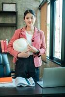 Professional woman engineer in office, holding helmet, with plans and laptop, showcasing modern workplace and career in engineering. photo