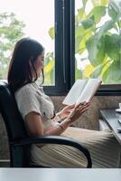 A woman sits in a comfortable chair reading a book by the window, with lush greenery visible outside. photo