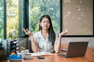 A young woman practices meditation at her desk in a modern office, surrounded by a laptop, notebook, and greenery outside the window. photo