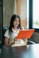 A woman enjoys reading an orange notebook while seated by a window in a contemporary office environment. photo