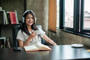 Smiling woman with headphones holding coffee cup, sitting at desk with open notebook in a bright office space. photo