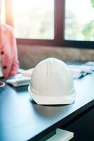 A white construction hard hat rests on an office desk, illuminated by sunlight through large windows, in a modern workspace setting. photo