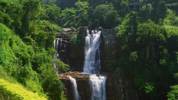 Water flows powerfully down a tiered waterfall surrounded by vibrant green vegetation in a sunny day, creating a breathtaking natural spectacle video