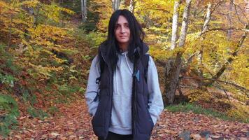 Happy confident Female hiker looking at camera walks through colorful autumn forest in surrounded by vibrant foliage and peaceful nature. Female traveler enjoys mindful walk connecting with nature, video