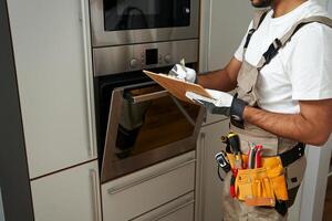Close up of service worker in uniform standing near oven and writing on clipboard in kitchen photo