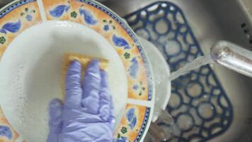 A gloved hand washes dirty dishes with a sponge in a sink under running water with suds video