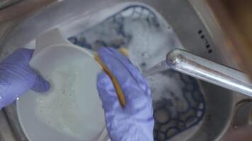 A hand in a blue glove washes a bowl with a sponge under running water in a sink with suds video