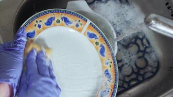 A hand in a blue glove washes a plate with a sponge in a sink with foam under running water video