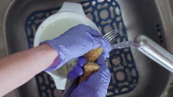 A womans hand in a blue glove washes a fork with a sponge in a sink under running water video