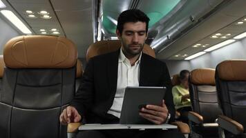A Businessman in a suit travels on airplane working on a tablet and speaking on the phone video