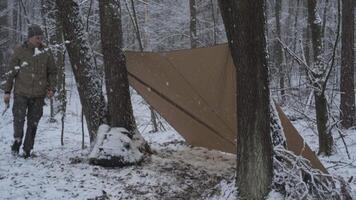A person sits in a snowy forest by a makeshift shelter, preparing gear while snowflakes fall around. The scene captures the quiet beauty of winter camping. video