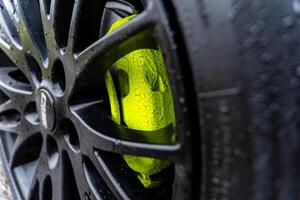 A striking lime-green brake caliper is partially visible through the black spokes of a car wheel, glistening with raindrops on a wet day, emphasizing the sleek design photo