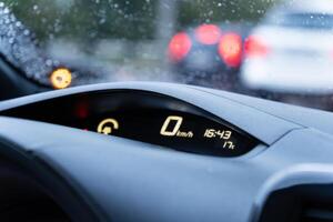 A car's dashboard features a speedometer reading zero while displaying the current time and temperature. Rain droplets are evident on the windshield, indicating wet weather photo