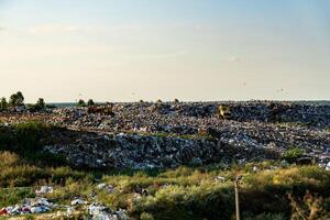 Heavy machinery works at a sprawling landfill filled with heaps of waste. The landscape is dotted with trees and a clear sky, indicating a warm, sunny day photo