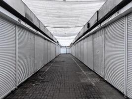 An empty marketplace features rows of closed stalls under a white canopy. The pavement is lined with tiles, and the atmosphere reflects a quiet afternoon in a commercial area photo