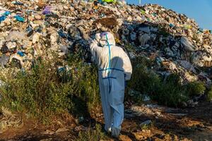 A person dressed in protective clothing walks through a landfill, examining the vast amount of waste. The bright sunlight illuminates heaps of garbage around them photo