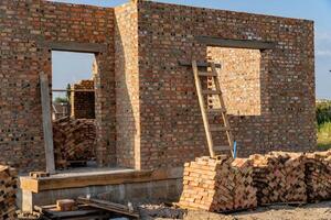 Construction workers are building a brick wall, which stands partially completed. A wooden ladder leans against the structure, and neatly stacked bricks nearby await use photo