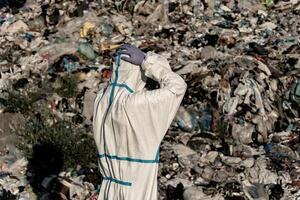 A worker wearing a protective suit examines a sprawling landfill overflowing with waste materials under bright daylight, highlighting the urgent issue of pollution and waste management photo